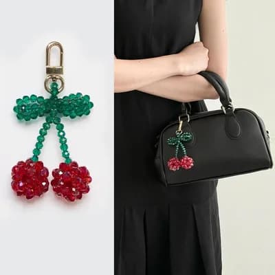 Two red cherries of faceted glass beads under a green bow, on a gold-tone clasp, and the same keychain on a black handbag