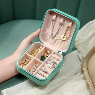 An open mint mini jewelry box held in a hand, with necklaces on the lid hooks and rings in the base