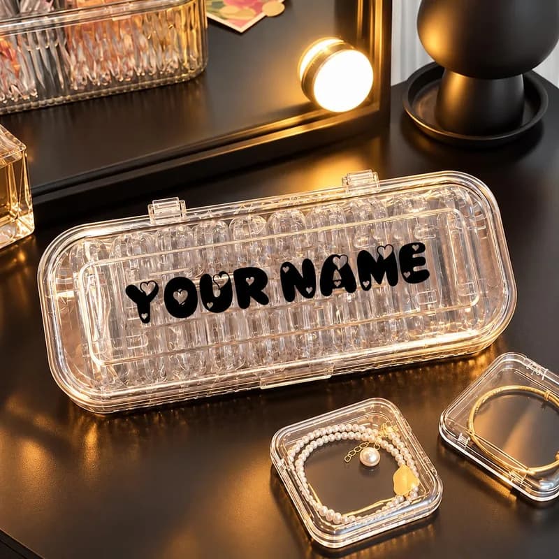 A clear ribbed case with “YOUR NAME” printed in black bubble letters on the lid, beside two small clear boxes holding bracelets