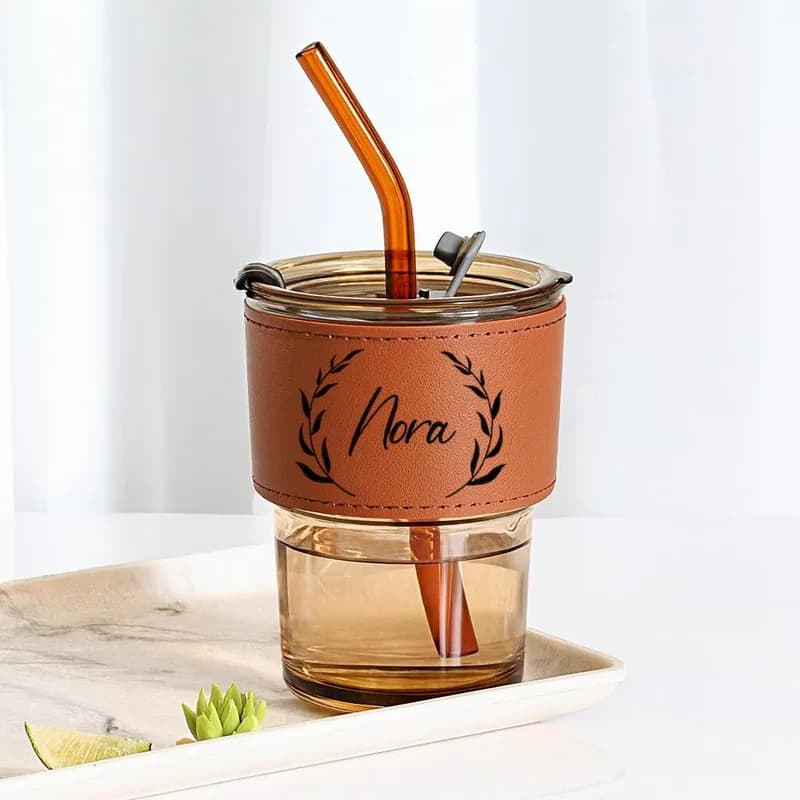 An amber glass tumbler with a straw and a brown sleeve engraved with the name Nora in a wreath