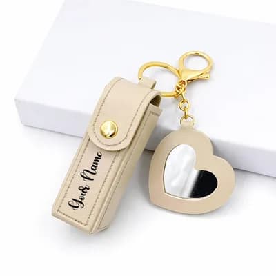 A cream lipstick case printed with “Your Name”, on a gold key ring with a cream heart mirror charm