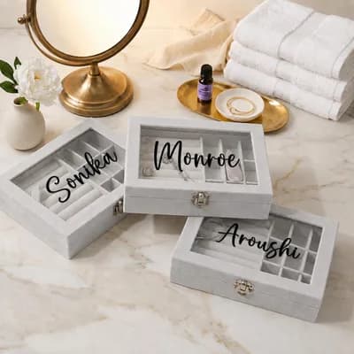 Three gray velvet boxes printed “Sonika”, “Monroe” and “Aroushi” in black script on their glass lids