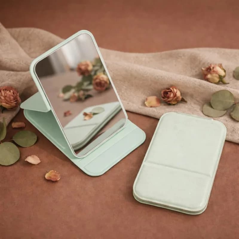 An open mint mirror on its stand beside its plain closed cover, among dried roses